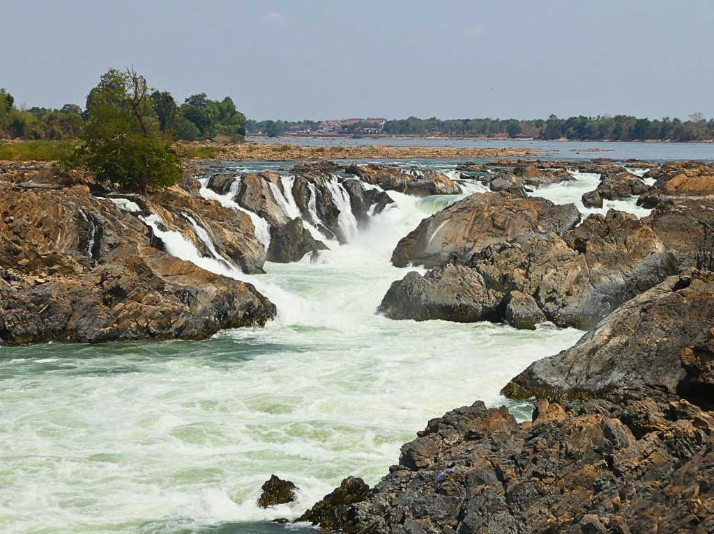 A scenic view of Khone Phapheng Falls in Laos, with cascading water rushing over rocky formations and lush greenery in the background.