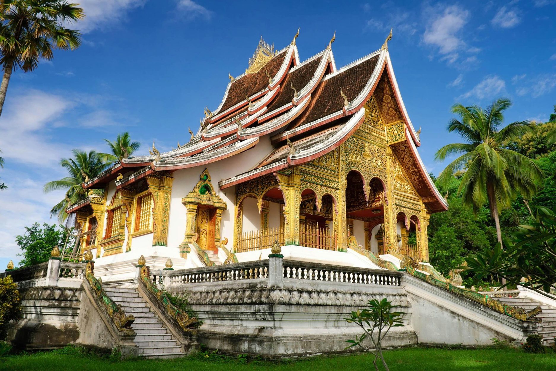 A beautifully ornate temple in Laos with intricate golden details and a blue sky in the background, surrounded by lush greenery and palm trees.