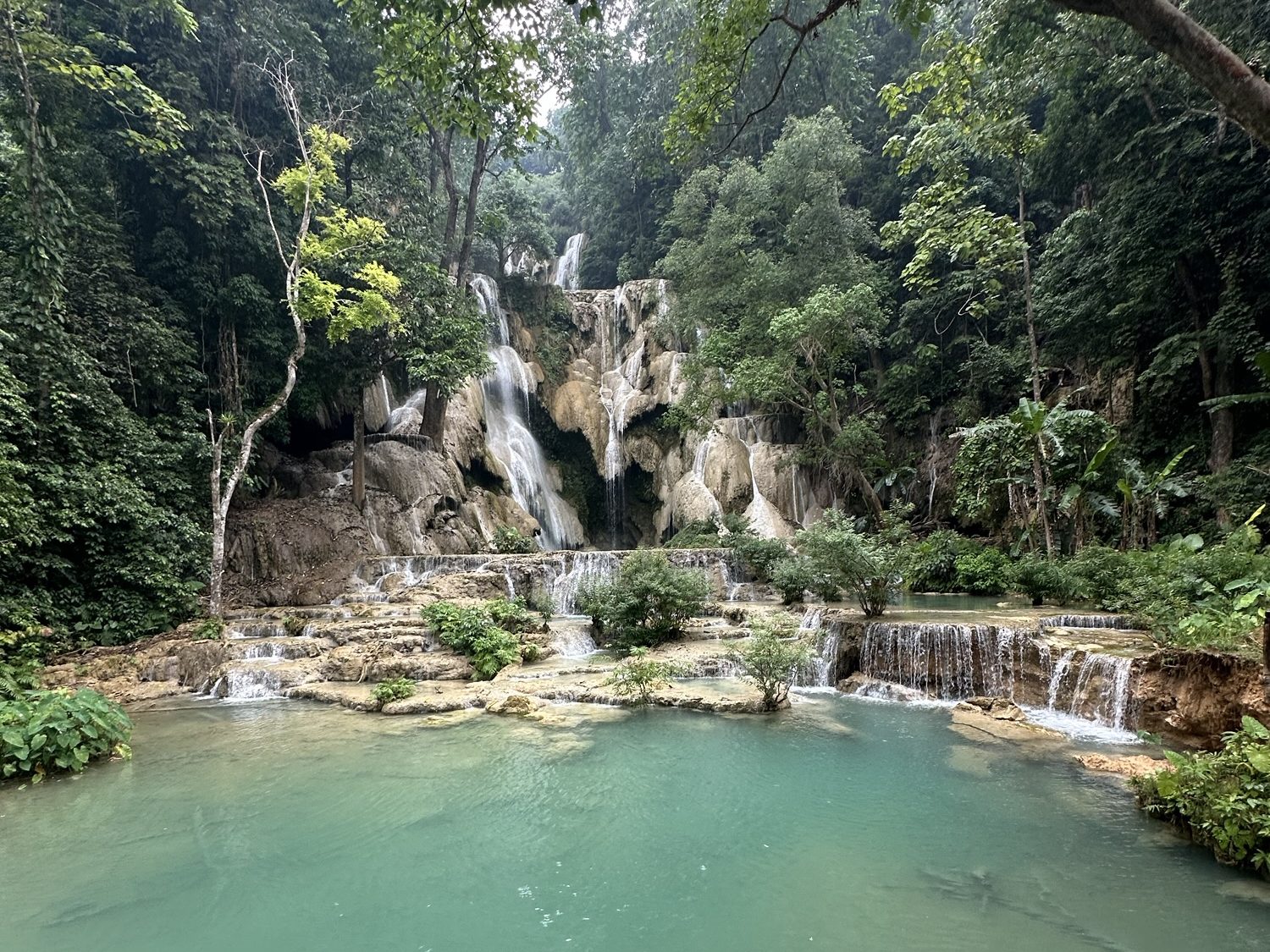 A picturesque waterfall cascading down rocky tiers surrounded by lush green forest and vibrant foliage, with crystal clear turquoise pools at its base.