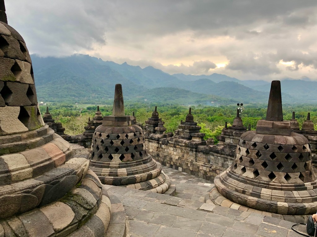 View of Borobudur Temple's stupas with lush hills and cloudy sky in the background.