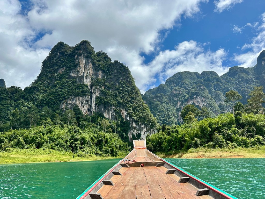 A scenic view from the bow of a boat navigating through a lake surrounded by lush green mountains under a partly cloudy sky.