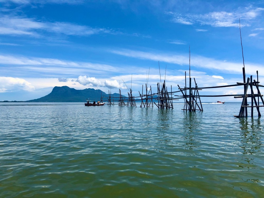 A serene view of calm waters with wooden fishing structures extending into the sea, set against a backdrop of lush hills under a bright blue sky with scattered clouds.