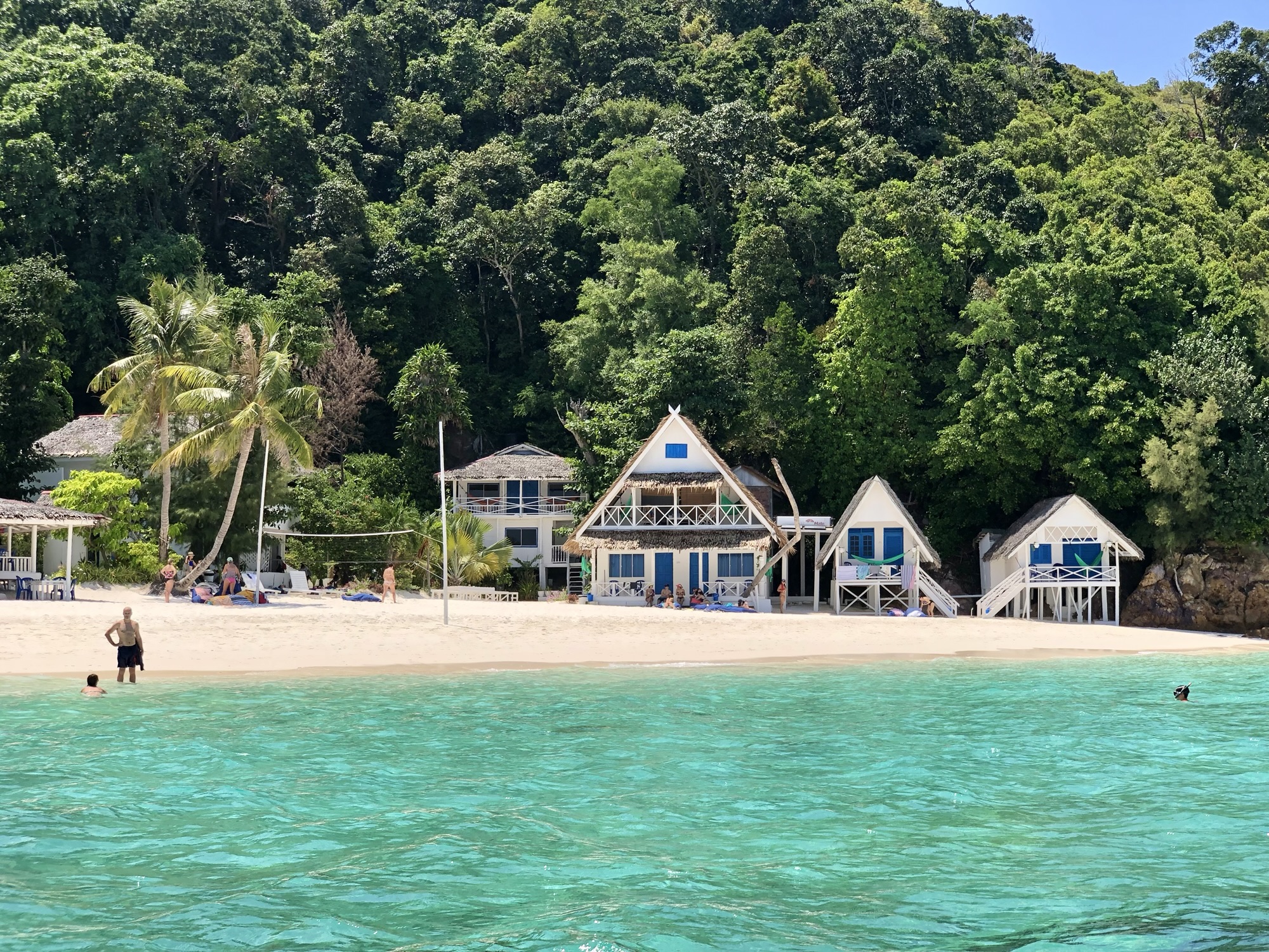 Tropical beach scene featuring turquoise water, a sandy shore, and large green trees in the background. Several people are relaxing on the beach and in the water near beachside cabins.