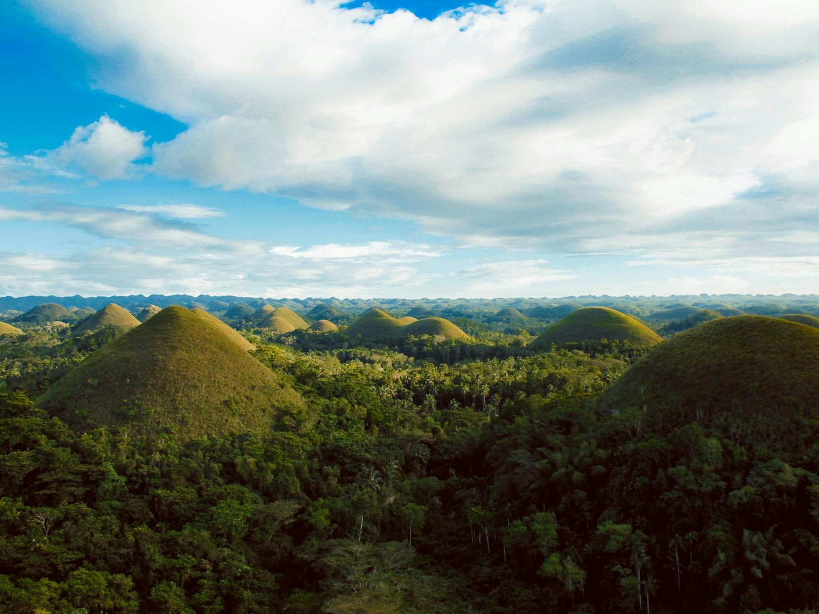 Aerial view of the Chocolate Hills in Bohol, Philippines, showcasing the distinct conical hills surrounded by lush greenery under a bright blue sky.