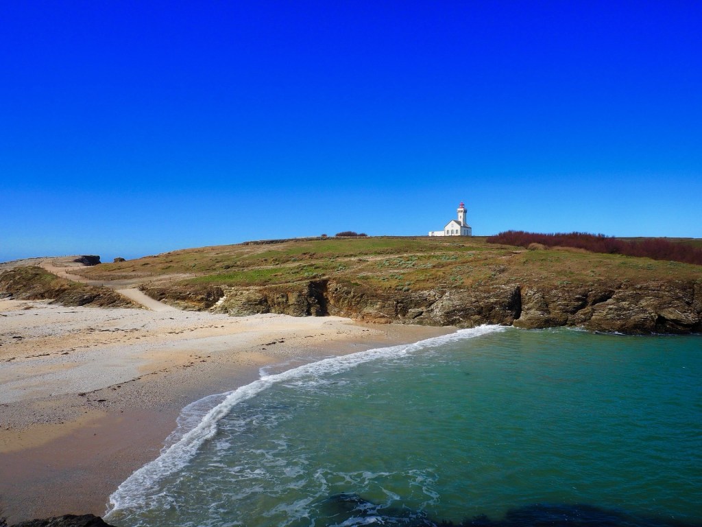 Vue d'une plage avec des eaux turquoise et un phare sur une colline, sous un ciel bleu clair.