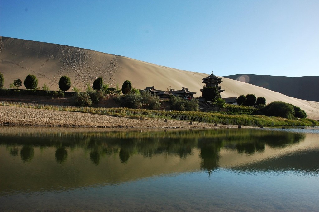 A serene landscape featuring a large sandy dune in the background, with a building surrounded by greenery and a calm body of water reflecting the scene.