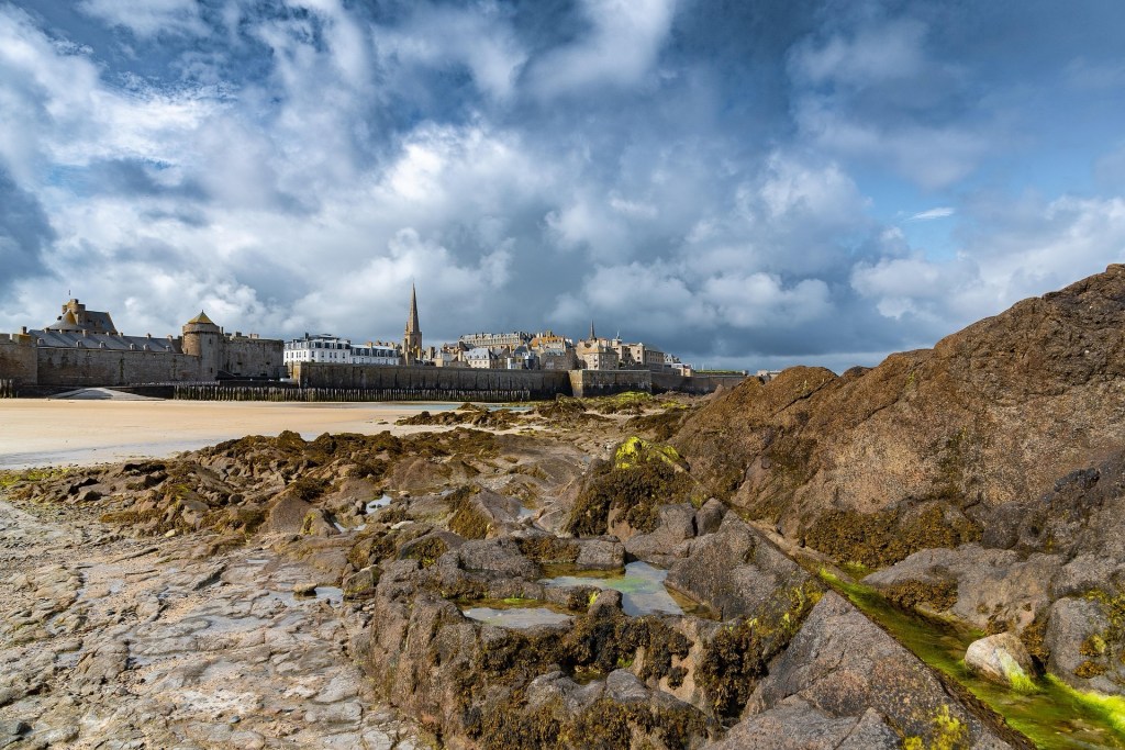 Vue de la ville de Saint-Malo avec des bâtiments historiques en arrière-plan, des rochers en premier plan, et un ciel nuageux.