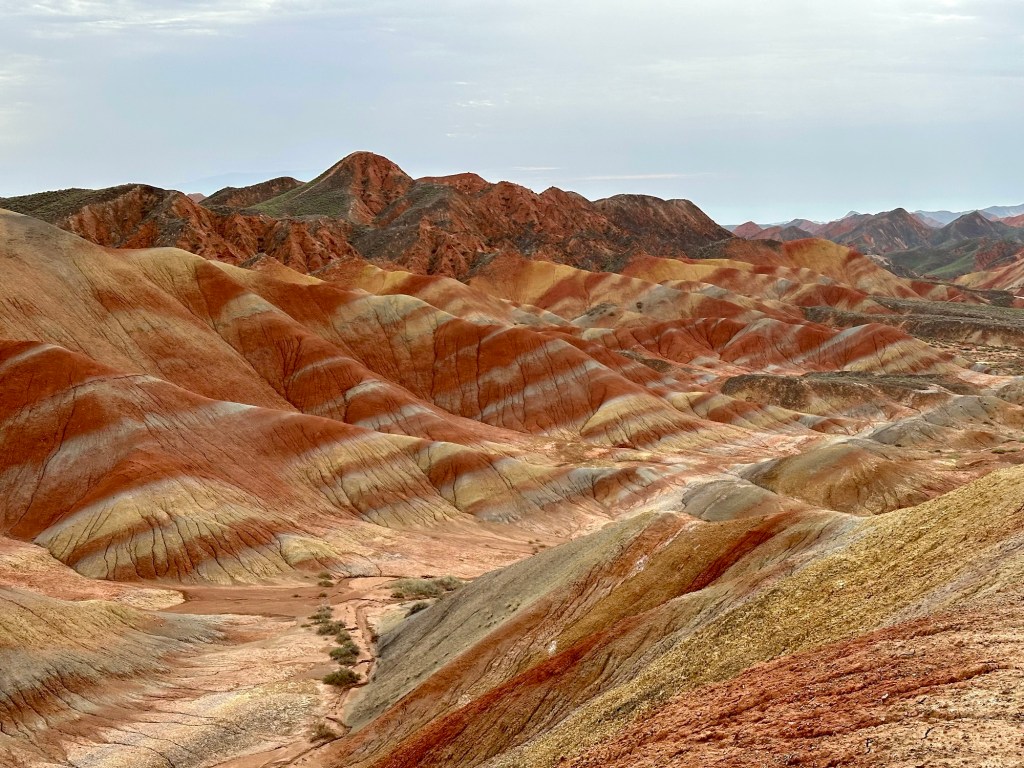 A panoramic view of the Danxia Landform, showcasing colorful, undulating hills with vibrant shades of red, orange, and yellow under a cloudy sky.