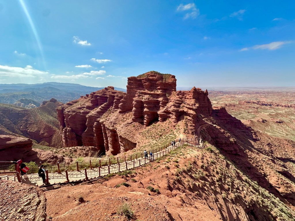A panoramic view of red rock formations in a canyon, with a pathway winding through the landscape and a clear blue sky above.