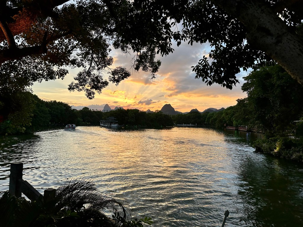 A serene view of a river at sunset, surrounded by lush trees and distant karst mountains, with a boat gently navigating the water.