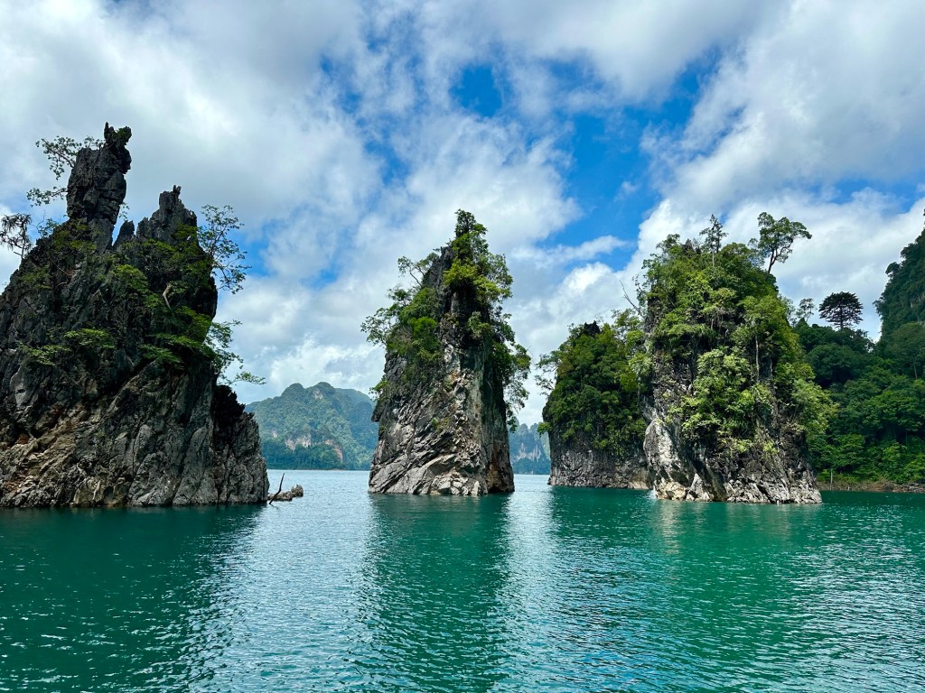 Scenic view of towering karst formations rising from the turquoise waters of Cheow Lan Lake, surrounded by lush greenery and cloudy blue skies.
