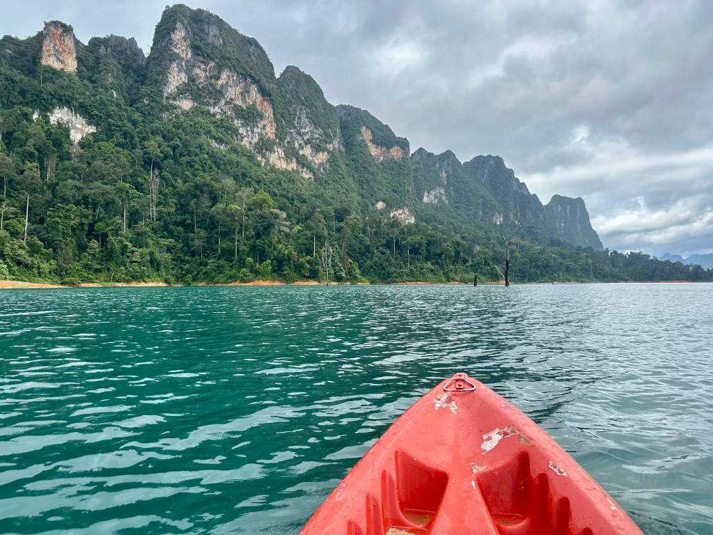A view from the front of a red kayak on a serene lake, surrounded by lush green forests and towering karst mountain formations under a cloudy sky.