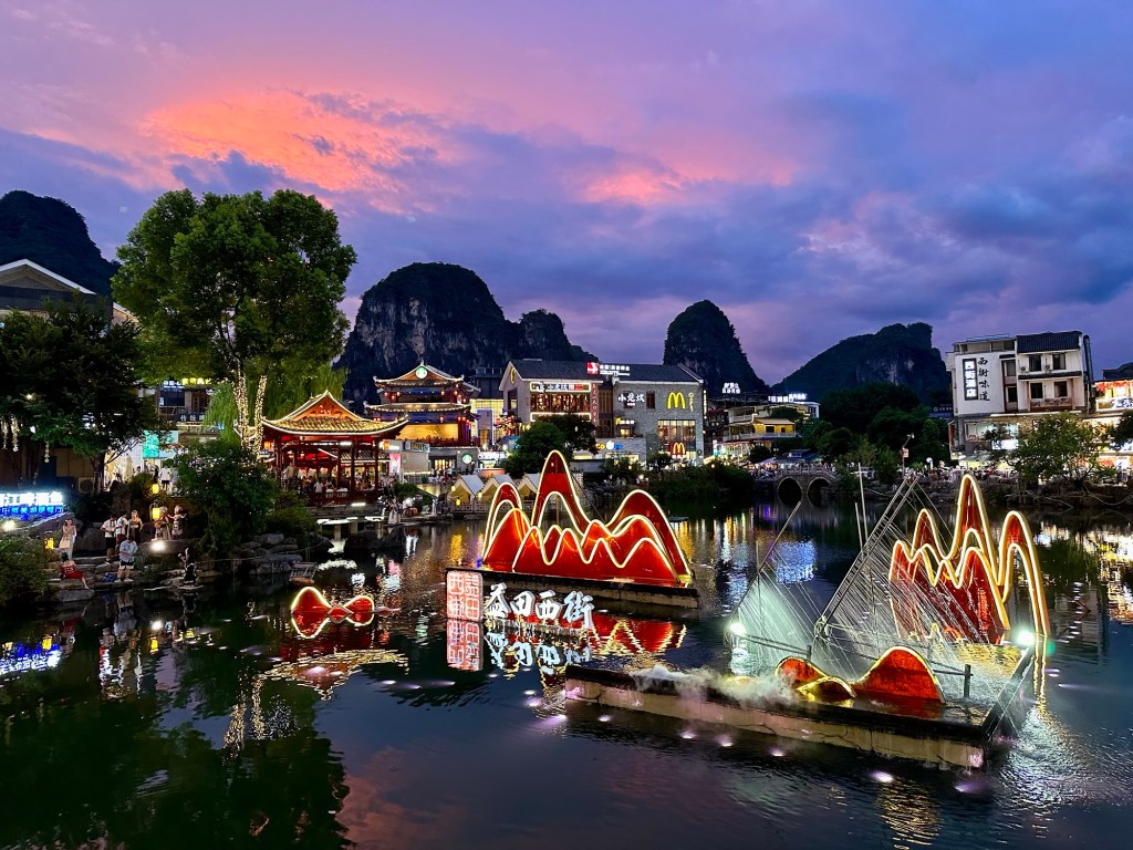 A picturesque view of a river in Yangshuo, China, illuminated by colorful lights at dusk, showcasing traditional architecture and karst mountains in the background.