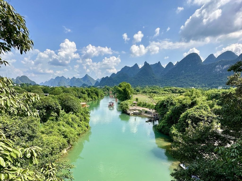 A picturesque scene showcasing a serene river winding through lush green vegetation, with dramatic karst mountains rising under a bright blue sky dotted with fluffy white clouds.
