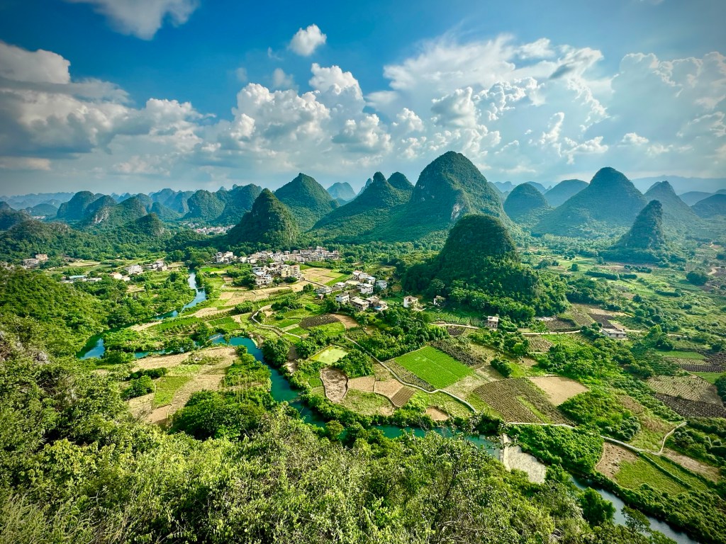 A panoramic view of the Guangxi landscape in China, featuring lush green fields, a winding river, and dramatic karst mountains under a clear blue sky.
