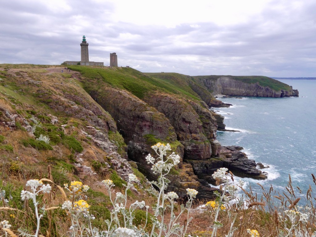 Vue des falaises côtières de Bretagne, avec un phare surplombant la mer. Des fleurs sauvages poussent sur le sol, tandis que des formations rocheuses bordent l'eau en contrebas.