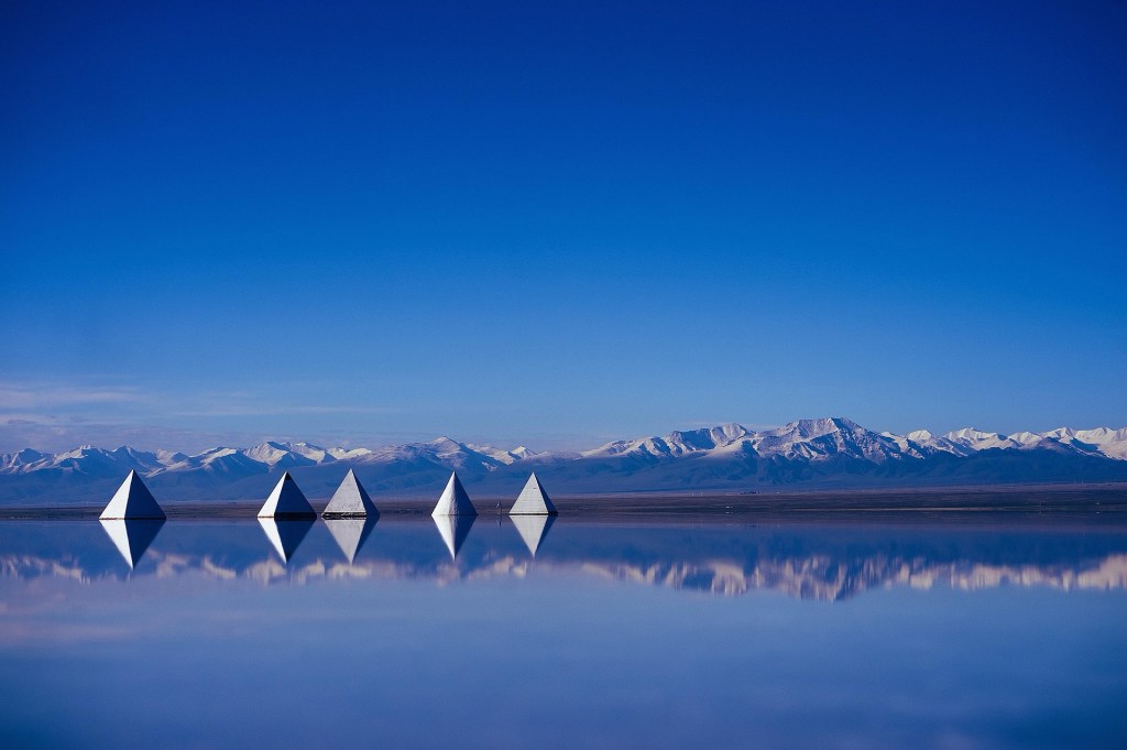 A serene landscape featuring pyramid-shaped structures reflecting in a tranquil salt lake, with snow-capped mountains and a clear blue sky in the background.