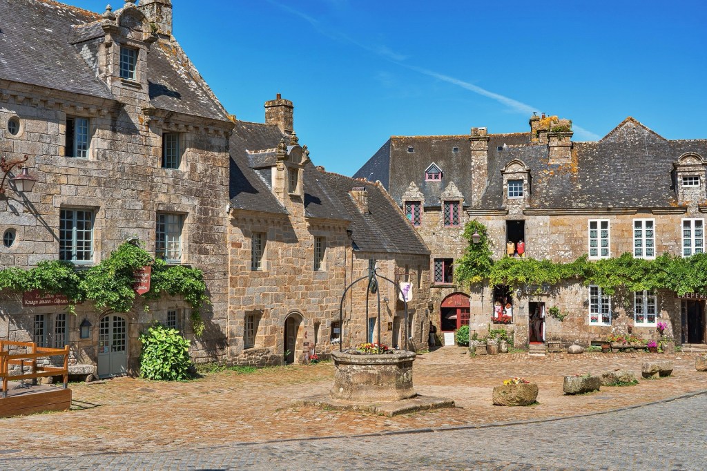 Vue d'une place pittoresque avec des maisons en pierre typiques de Bretagne, entourées de verdure et de fleurs colorées sous un ciel bleu.